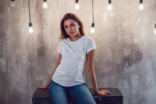 Sexy vrouw in een witte T-shirt op een grijze achtergrond met lampen. Mock-up. — Stockfoto