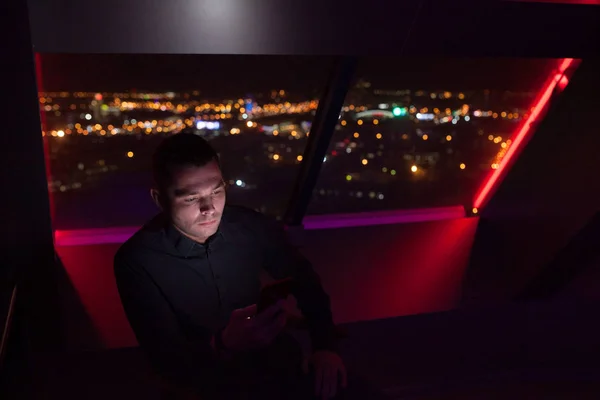 Empresário usa um smartphone sentado à janela no escritório em um fundo de uma cidade noturna . — Fotografia de Stock