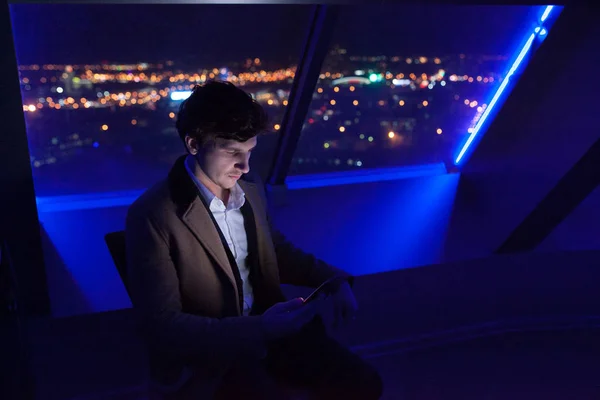 ビジネスマンは、夜の街の背景のオフィスの窓辺に座っているスマート フォンを使用してください。. — ストック写真