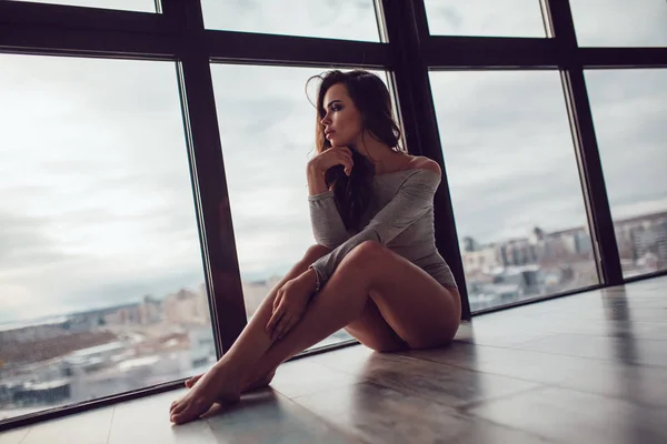 Mujer sexy en traje de cuerpo gris se sienta en el suelo cerca de la ventana panorámica . —  Fotos de Stock