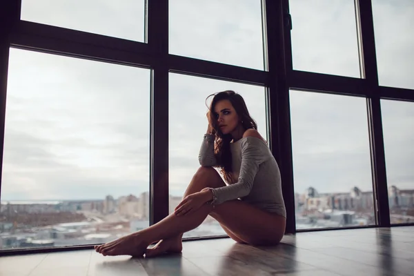 Sexy Frau im grauen Body sitzt auf dem Boden neben dem Panoramafenster. — Stockfoto