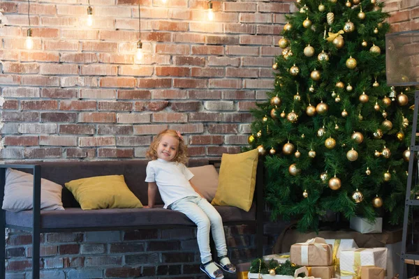 Jolie petite fille dans un t-shirt blanc est assis sur le canapé près du sapin de Noël. Maquette . — Photo