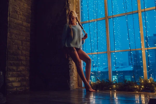 Sexy blond dziewczyna w sweter stoi w pobliżu okna. — Zdjęcie stockowe