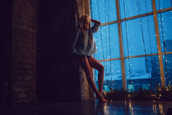 Menina loira sexy em uma camisola fica perto de uma janela . — Fotografia de Stock