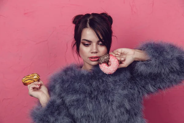 Menina bonita em um casaco de pele com donuts em mãos posando em um fundo rosa . — Fotografia de Stock