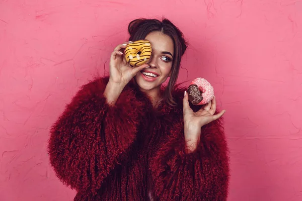 Fille dans un manteau de fourrure lumineux avec des beignets dans les mains posant sur un fond rose . — Photo