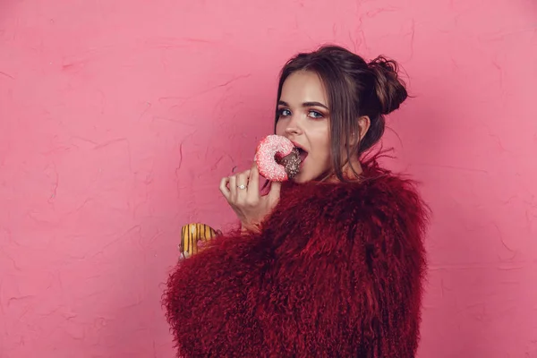 Menina em um casaco de pele brilhante com donuts em mãos posando em um fundo rosa . — Fotografia de Stock