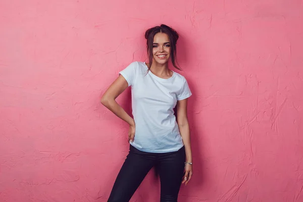 Una atractiva mujer con una camiseta blanca está de pie sobre un fondo rosa. Mock-up. —  Fotos de Stock