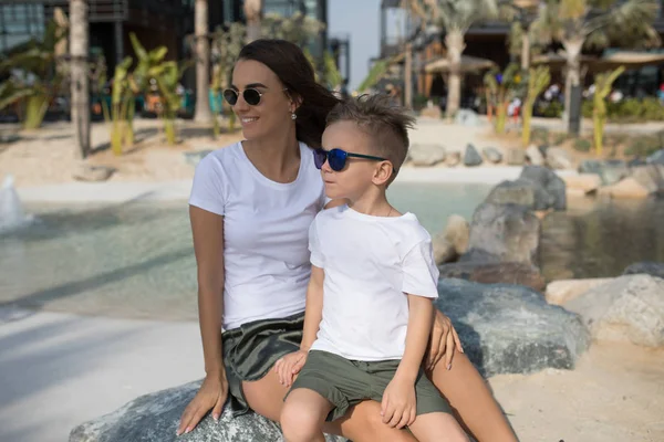 Kadın oğlunu sahilde oturan beyaz T-shirt ile.