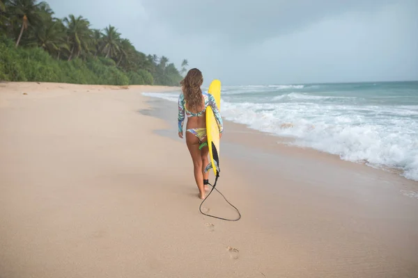Bakifrån av en kvinna surfare promenader längs stranden med en styrelse i händer. — Stockfoto