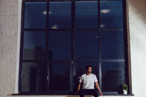 En afroamerikan i vit t-shirt som sitter på ett fönster. Modellering. — Stockfoto