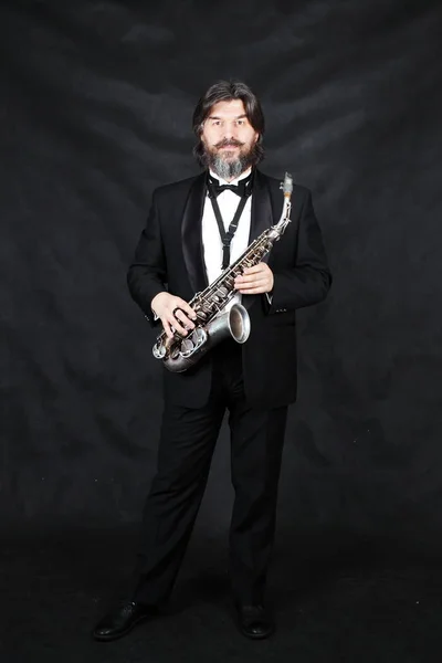 Full length profile shot of male artist musician in a classic black suit, tailcoat, statuesque in a bow tie with a beard plays music on a gold saxophone.black background