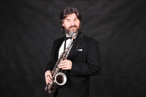 A male artist musician in a classic black suit, tailcoat, statuesque in a bow tie with a beard plays music on a gold saxophone.black background