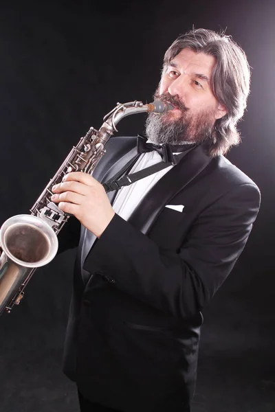 A male artist musician in a classic black suit, tailcoat, statuesque in a bow tie with a beard plays music on a gold saxophone.Playing an instrument against a white light source.black background