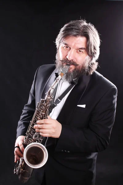 A male artist musician in a classic black suit, tailcoat, statuesque in a bow tie with a beard plays music on a gold saxophone.Playing an instrument against a white light source.black background