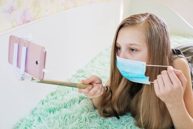 Kız koruyucu bir maske takıyor, evde eğleniyor, küresel bulaşıcı koronavirüs karantinası sırasında yüz hareketleri konseptinin yardımıyla telefonla fotoğraf çekiyor.