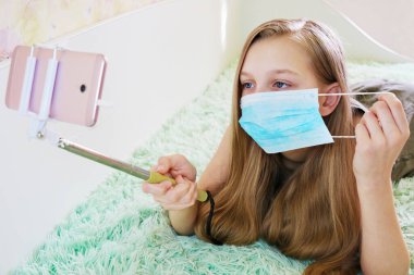 Kız koruyucu bir maske takıyor, evde eğleniyor, küresel bulaşıcı koronavirüs karantinası sırasında yüz hareketleri konseptinin yardımıyla telefonla fotoğraf çekiyor.