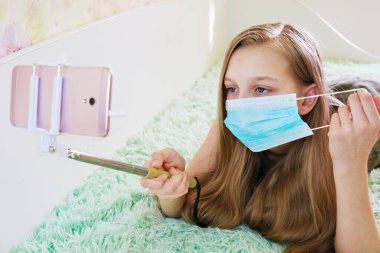Kız koruyucu bir maske takıyor, evde eğleniyor, küresel bulaşıcı koronavirüs karantinası sırasında yüz hareketleri konseptinin yardımıyla telefonla fotoğraf çekiyor.