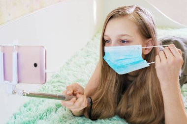 Kız koruyucu bir maske takıyor, evde eğleniyor, küresel bulaşıcı koronavirüs karantinası sırasında yüz hareketleri konseptinin yardımıyla telefonla fotoğraf çekiyor.