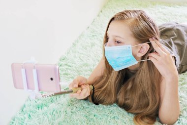 Kız koruyucu bir maske takıyor, evde eğleniyor, küresel bulaşıcı koronavirüs karantinası sırasında yüz hareketleri konseptinin yardımıyla telefonla fotoğraf çekiyor.
