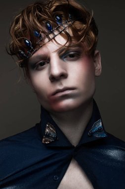 Bir Kral yaratıcı makyajlı suretinde bir genç adam. Kızıl saçlı ve mavi taş kafa ile taç. Parlayan cilt. Yüz güzelliği. Stüdyoda çekilmiş fotoğrafı.