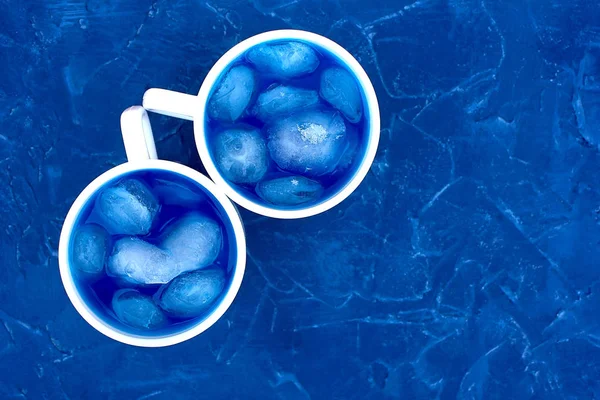 Blue royal color background and two white cups with blue water with ice cubes, a cooling drink