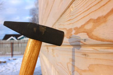 Ahşap bir duvarın arka planında demir bir çekiç tahtaya çivi çakar. Arka planda çit, ev ve gökyüzü.