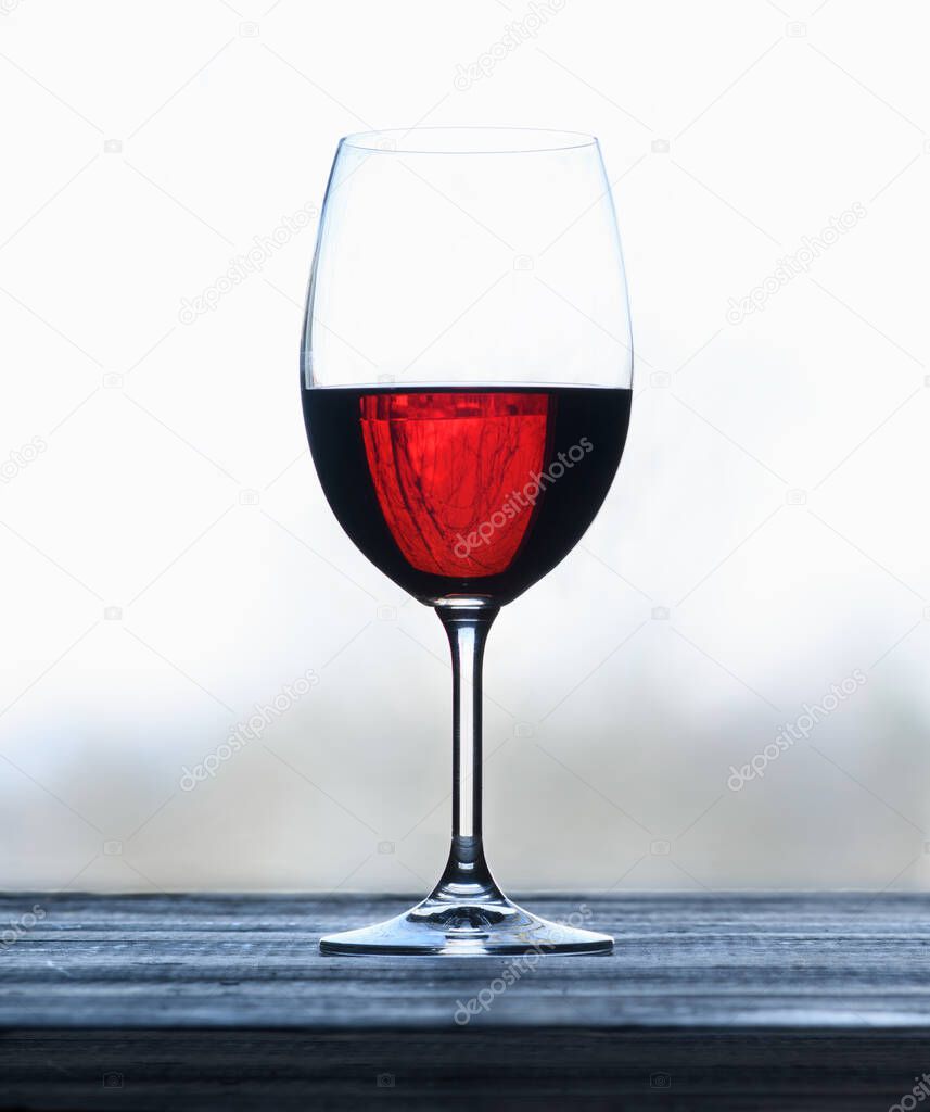 Glass with red wine on a wooden surface against a light blurred background.  Selective focus. The wine glass reflects the branches of a tree.