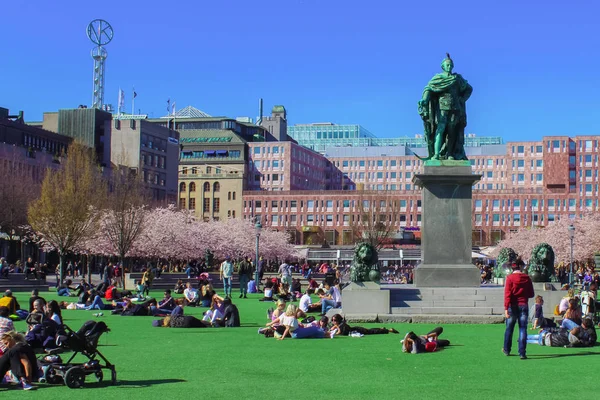Stockholm, Svédország - 2019. április 28.: Családok ülnek zöld füvön XIII. Károly svéd király szobra mellett a Kungstradgarden-ben a rózsaszín cseresznyevirágzás idején tavasszal. Az emberek élvezik a tavaszt. Családtag. — Stock Fotó