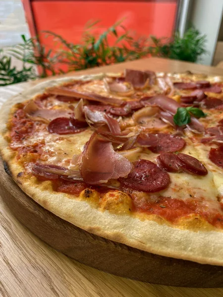 ピッツェリア内の赤い壁の背景と緑の植物と木製のテーブルの上に熱いイタリアのピザを閉じます サラミと揚げフラットブレッド チーズ 揚げハムと新鮮な緑のバジル 縦型携帯写真 — ストック写真