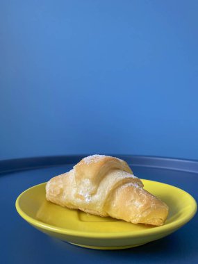 Mavi masanın üzerine mavi modaya uygun arka planda beyaz şeker tozu serpiştirilmiş taze kruvasan serpiştirilmiş sarı tabak. Cep telefonu fotoğrafı. Kahvaltı konsepti