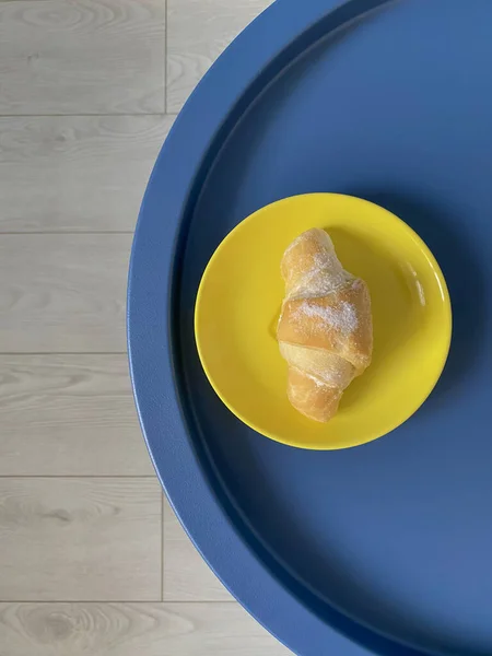 stock image Top view on yellow plate with fresh croissant on round blue table with white wooden floor as background. Mobile photo. Breakfast concept