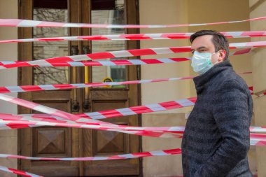 Coronavirus Covid-19 pandemisi sırasında tek kullanımlık koruyucu maske takan yakışıklı Avrupalı genç bir adam. Kapalı restoran veya dükkan kapısının yanında kırmızı bantlı..