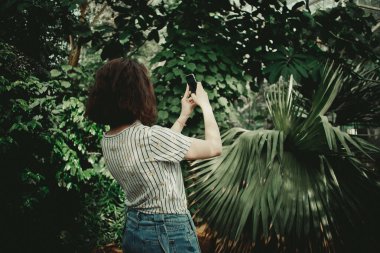 Bir kız telefonda büyük bir serada ağaçların ve bitkilerin fotoğrafını çekiyor. Palmiye ağaçları, muzlar, çalılar ve güzel fotoğraflar..