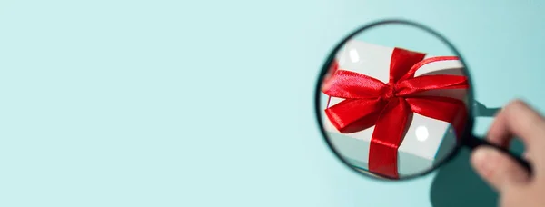Una confezione regalo bianca con un fiocco rosso ingrandito attraverso una lente d'ingrandimento tenuta a mano. Sullo sfondo menta — Foto Stock