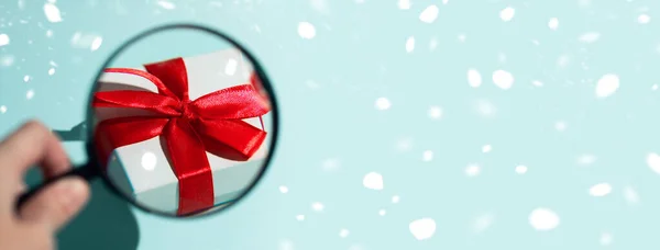 Un coffret cadeau blanc avec un arc rouge magnifié à travers une loupe tenue à la main. Sur fond de menthe — Photo
