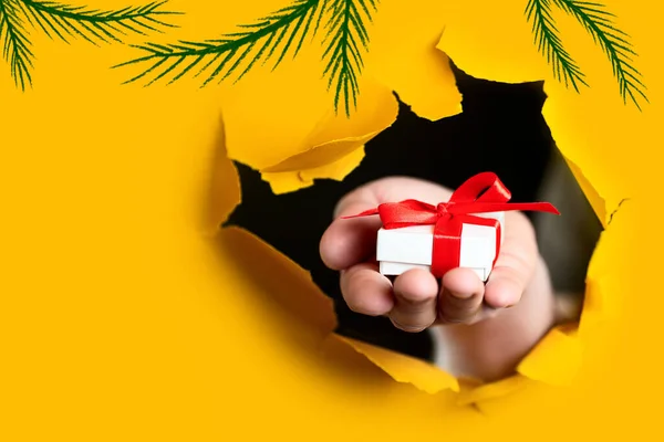 Un regalo con un lazo rojo en la mano emerge de un agujero irregular en el fondo de papel amarillo — Foto de Stock