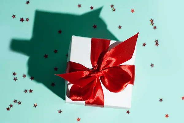 Weiße Geschenkschachtel mit roter Schleife und einer Sternenverstreuung auf mintfarbenem Hintergrund. der Schatten ist wie ein Haus. — Stockfoto
