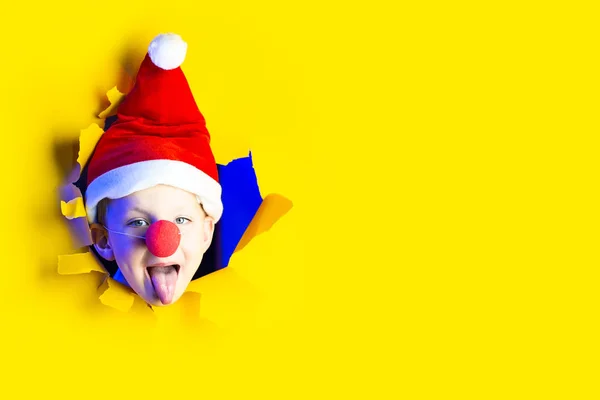 En dreng i en julemandshat og en klovns næse viser sin tunge fra et hul i det gule papir. Reklameplads . - Stock-foto