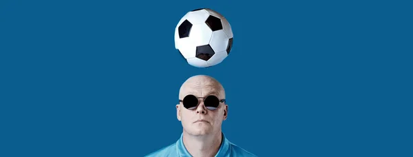 Calvo hombre brutal en gafas redondas oscuras con reflejos. Un balón de fútbol sobrevolaba. Sobre un fondo azul . — Foto de Stock