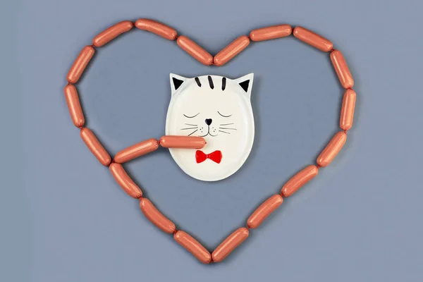 Talerz kota zjada kiełbaski rozłożone przez serce wokół niego. Na szarym tle. Koncepcja Walentynek — Zdjęcie stockowe