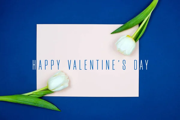 Blank różowy arkusz papieru i świeże kwiaty tulipan z zielonymi liśćmi na niebieskim tle, widok z góry — Zdjęcie stockowe