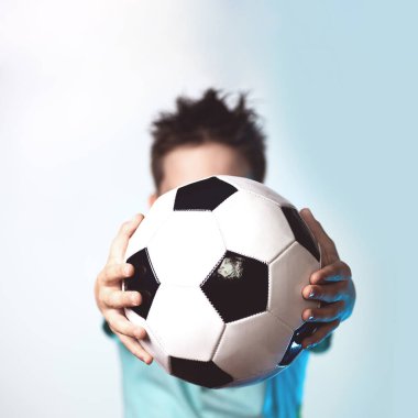 elinde bir futbol topu tutan mavi bir t-shirt bir çocuk kafasını mavi bir arka planda gizler
