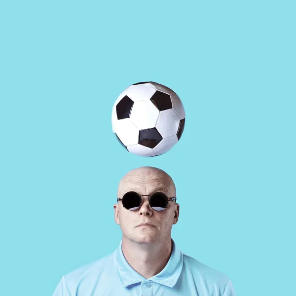 Calvo hombre brutal en gafas redondas oscuras con reflejos. Un balón de fútbol sobrevolaba. Sobre un fondo azul . — Foto de Stock