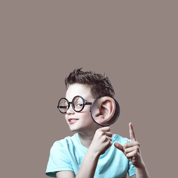 一个穿着蓝色T恤和眼镜的男孩给他的耳朵带来了放大镜 是米色背景的关于远程学习问题的概念 — 图库照片