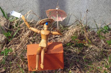 A wooden man sits on a brick under an umbrella after the quarantine is lifted. He threw off his mask and began to sunbathe. Concept on the topic of the pandemic clipart