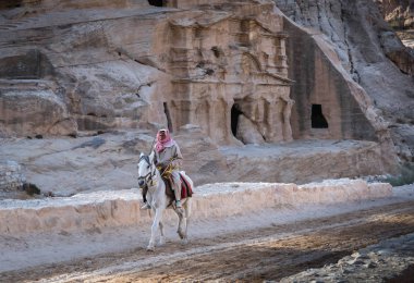 Petra, Jordan - November 28 2017: Bedouin man riding a white horse with ancient building carved in the rock in the background nearby the archaeological area of the Treasury, UNESCO World Heritage Site