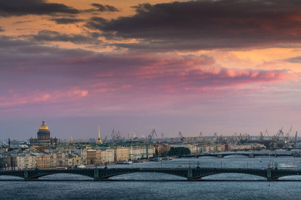 Красивый летний вид на Неву и Санкт-Петербург с высоты заката солнца: Троицкий мост, Зимний дворец, Исаакиевский собор и портовые краны вдалеке
