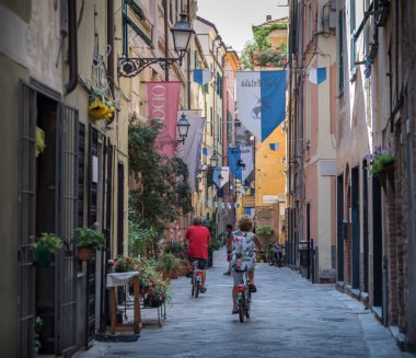 Albenga, Savona, İtalya - 21 Temmuz 2018: İtalyan ortaçağ eski şehir sokağında erkek ve kadın bisikleti, tipik Liguria renkli cepheleri, dar sokakları ve geleneksel palio bayrağı