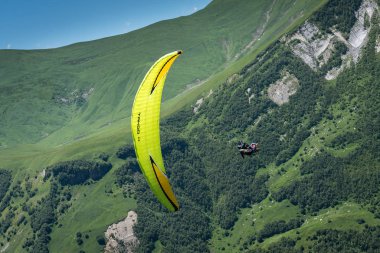 Gudauri, Caucasus, Tbilisi, Georgia - July 2019: Paragliding in the Caucasian mountains at Jvari Pass nearby Georgia Russia Friendship Monument. Yellow wing, green grass, meadows, fields, and forests 